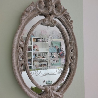 Espelho oval com moldura ornamentada cinza desgastado refletindo prateleira com objetos