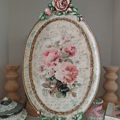 Placa decorativa oval com flores cor-de-rosa e moldura dourada em ambiente com castiçais e potes.