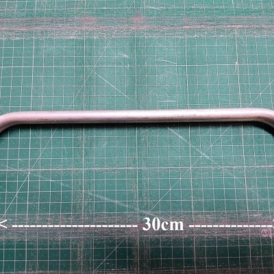 Barra metálica prateada com extremidades douradas sobre superfície verde com grelha e indicação 30cm