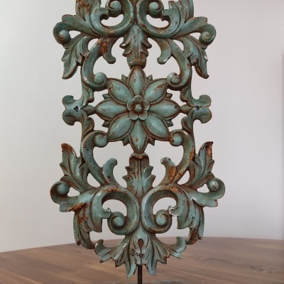 Ornamento decorativo verde-azulado com padrão floral em metal sobre mesa de madeira