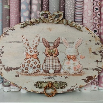 Placa decorativa oval com três coelhos pintados em padrões florais e xadrez com laços, encostada em rolos de tecido coloridos e estampados em tons rosa e cinza