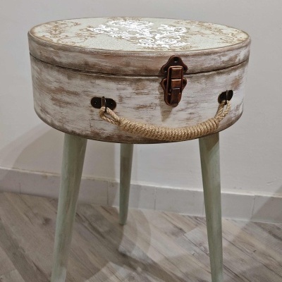 Mesa auxiliar redonda de madeira com tampa decorada, fecho metálico e alça de corda, apoiada em três pernas verdes.