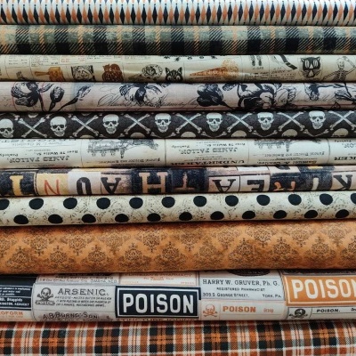 Rolos de tecido com padrões variados como xadrez, caveiras, bolinhas e textos 'ARSENIC', 'POISON' em fundo bege e castanho.