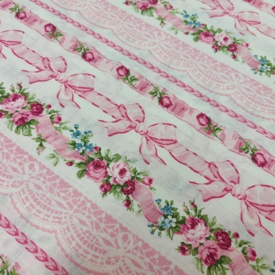 Pano branco com padrão floral em rosa e verde, laços e rendas cor de rosa.