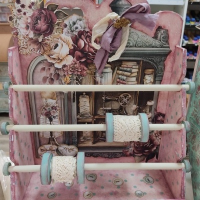 Suporte de madeira decorativo para fitas com fitas rendadas e decoração floral em tons rosa, verde e azul.