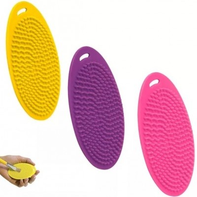 Três escovas de silicone oval coloridas amarelo, roxo e rosa com textura lisa e buracos para pendurar.