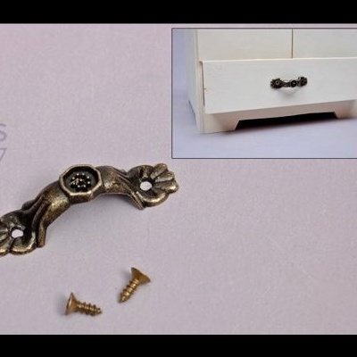 Puxador de móvel metálico com motivos florais e dois parafusos dourados têm gaveta branca como fundo.