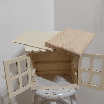 Casinha decorativa de madeira clara com portas duplas abertas sobre cadeira branca.