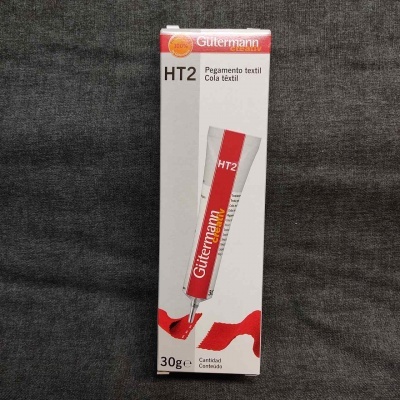 Embalagem de cola têxtil Gutermann HT2 vermelha e branca sobre fundo cinza.