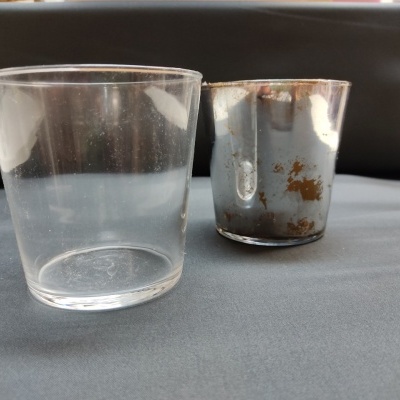 Dois copos pequenos, um de vidro transparente e outro metálico com manchas