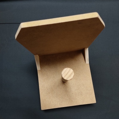 Objeto de madeira clara com base quadrada e placa vertical sobre fundo escuro