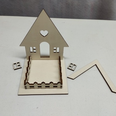 Peça desmontada de madeira clara em forma de casa com cerca e telhado separado