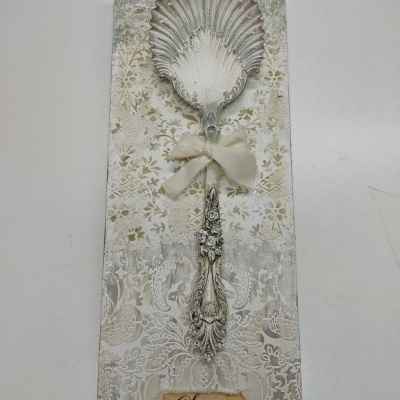 Utensílio decorativo em forma de colher grande com padrão de concha e fita creme em base decorativa com etiqueta Spoon.