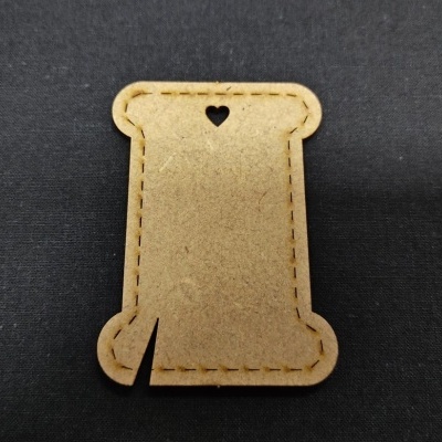 Acessório em forma de carretel de linha em material castanho claro com recorte em forma de coração sobre fundo preto