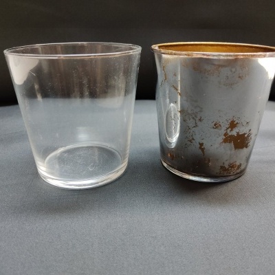 Dois copos pequenos, um de vidro transparente e outro metálico envelhecido, sobre superfície cinza