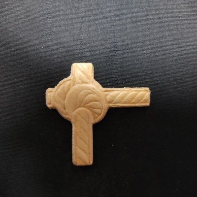 Peça de madeira clara em forma de cruz celta entalhada sobre fundo preto.