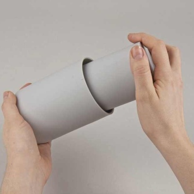 Caixa cilíndrica cinza fosca sendo encaixada por duas mãos sobre fundo cinza claro