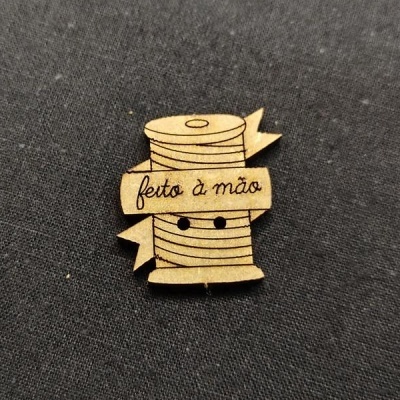 Botão de madeira com design de rolo de linha e texto 'feito à mão' sobre tecido preto