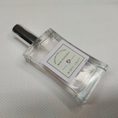 Frasco de perfume transparente com tampa preta e rótulo branco sobre fundo branco com pontos cor de rosa