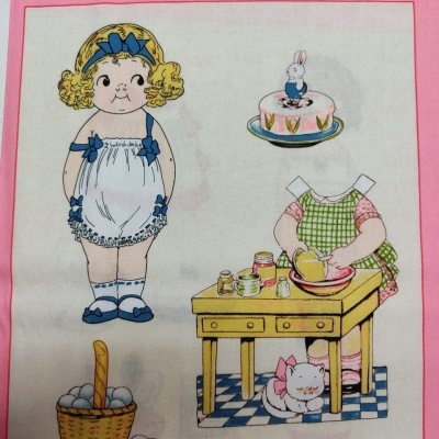 Lenço com desenho colorido de boneca, bolo, figura sem cabeça a cozinhar, cesta de ovos e gato junto a mesa