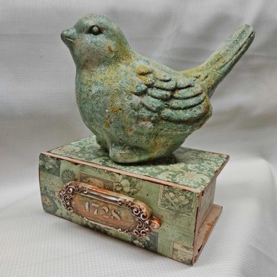 Estatueta de pássaro verde envelhecido sobre caixa decorativa com placa '1728'.