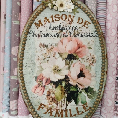 Placa oval decorativa em metal com flores e texto em francês, rodeada de tecidos coloridos com padrões variados