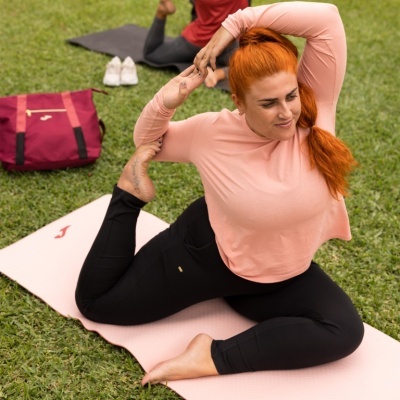 Mulher a fazer yoga num tapete creme com logótipo vermelho no relvado, mochila vinho e tênis brancos ao fundo