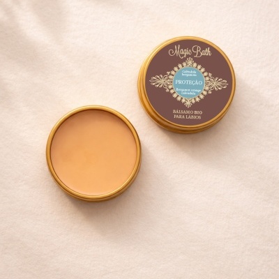 Bálsamo para lábios em embalagem redonda dourada com rótulo castanho e azul, e bálsamo aberto cor de pêssego em fundo bege.