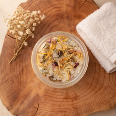 Frasco de sais de banho com flores secas sobre base de madeira com toalha branca e flores secas ao lado.