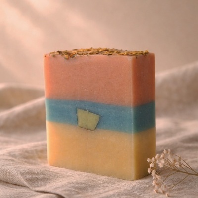 Sabonete artesanal em camadas rosa, azul e amarelo com sementes no topo e flores secas ao lado