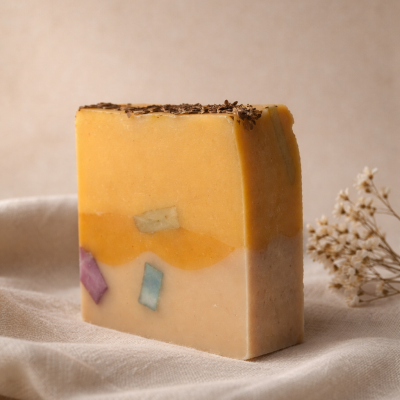 Sabonete artesanal amarelo e bege com cubos coloridos e sementes no topo, sobre tecido bege.