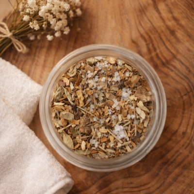 Frasco com mistura de ervas secas, toalha branca e flores pequenas sobre madeira