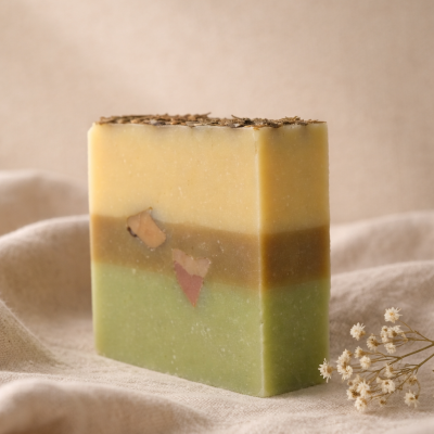 Sabonete artesanal retangular com camadas verde, castanho e amarelo e sementes secas no topo sobre tecido bege