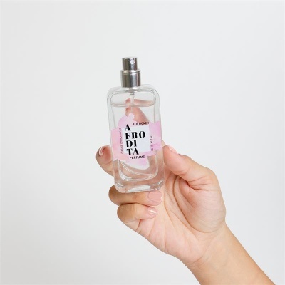 Frasco de perfume transparente com rótulo e líquido rosa sendo segurado por uma mão