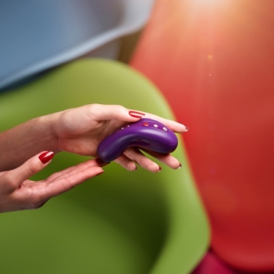 Dispositivo eletrônico roxo sendo segurado por mãos femininas com unha vermelha, poltrona verde ao fundo.