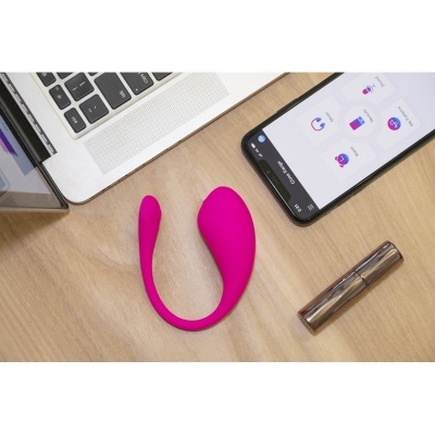 Dispositivo de silicone rosa para uso íntimo ao lado de portátil, smartphone e objeto metálico numa mesa de madeira