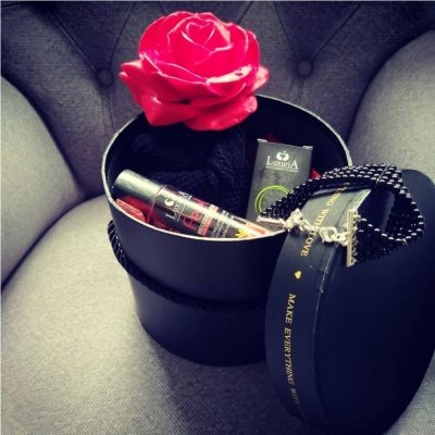 Caixa preta redonda com rosa vermelha, frasco, caixa verde e acessório preto numa cadeira cinza