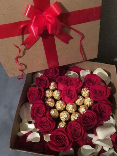 Caixa castanha com laço vermelho contendo rosas vermelhas, pétalas brancas e bombons dourados Ferrero Rocher