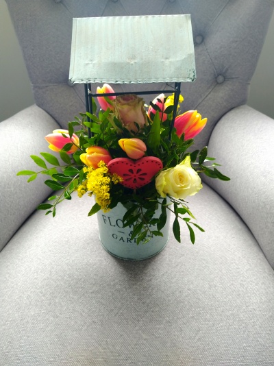 Arranjo floral em vaso metálico com capa metálica, flores coloridas e coração decorativo, sobre cadeira cinza.