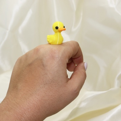 Anel infantil amarelo em forma de pato sobre a mão humana, fundo branco.