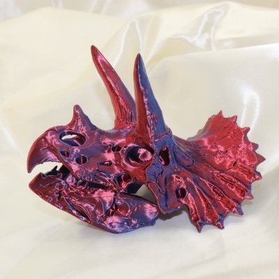 Escultura metálica vermelha e azul de crânio de dinossauro sobre tecido branco.
