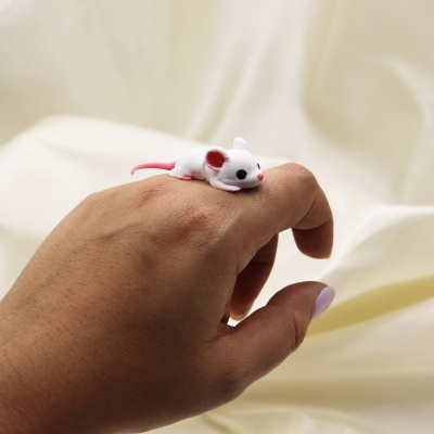 Anel com figura de rato branco e rosa em dedo sobre fundo bege.