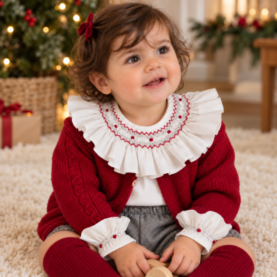 Bebé vestida com camisola vermelha, gola branca bordada, calções cinza e meias vermelhas, sentada em carpete com decoração de Natal ao fundo.