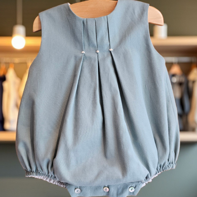 Body de bebé azul claro com pregas e botões, pendurado em cabide de madeira