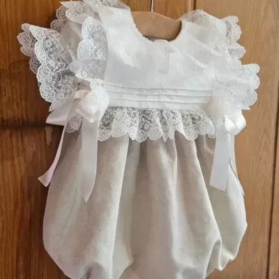 Macacão de bebé bege e branco com renda e folhos pendurado num cabide de madeira