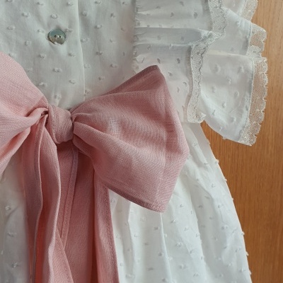 Vestido branco de criança com laço rosa e folhos nas mangas, pendurado num cabide de madeira.