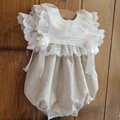 Macacão de bebé bege com renda branca e fitas sentado numa cabide de madeira na porta