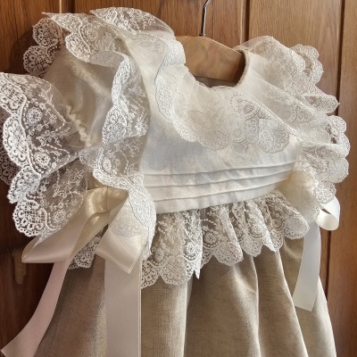 Vestido bege claro para bebé com gola de renda branca e laços de cetim em cabide