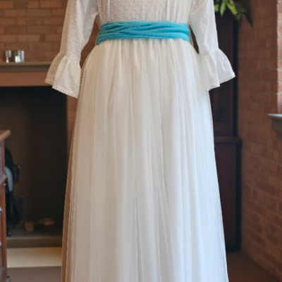 Vestido branco comprido com cinto azul claro em manequim sobre fundo de parede de tijolos