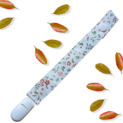 Prendedor de chupeta floral branco com clip e fecho branco rodeado por folhas secas.